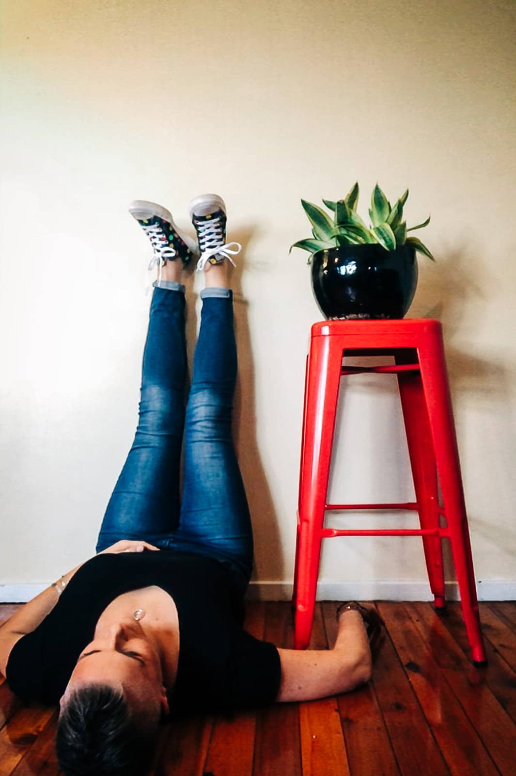 Lying on the floor with legs up on the wall. A red stool with a plant in a black pot is next to the person