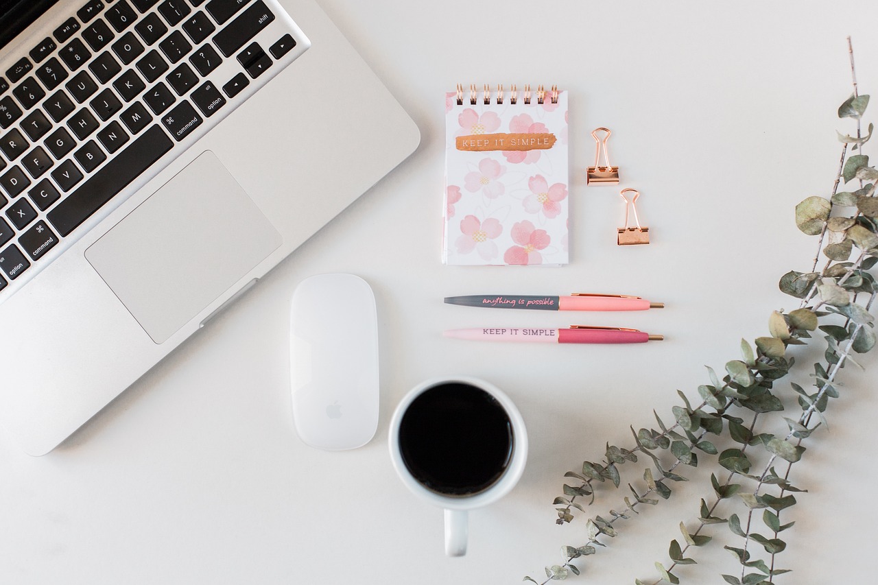 Top down view of Macbook laptop with Apple mouse. Black coffee in a white cup. A pink and white spiral bound notebook with pink pens and pink bulldog clips, and a sprig of eucalyptus leaves.