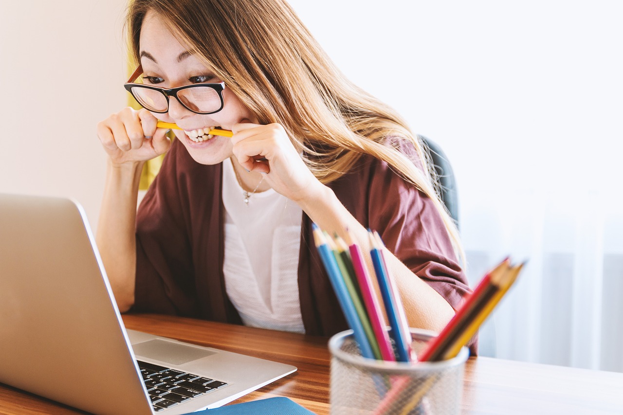 A woman with long straight hair and glasses leaning over a Macbook laptop, holding a yellow pencil between her teeth. She has a frustrated look on her face. On the desk is a jar with more coloured pencils standing up in it